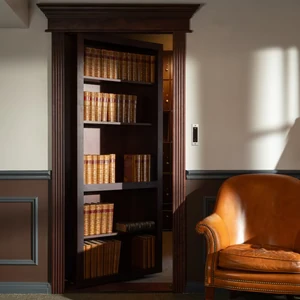 Dark wood bookcase swings open like a hidden door in a paneled room, beside a tan leather chair.