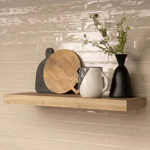 Light wood floating shelf on a tiled wall, styled with cutting boards, a pitcher, and a vase.