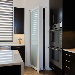 Modern kitchen with black cabinetry and stainless steel ovens, featuring a white hidden door with frosted glass panel.