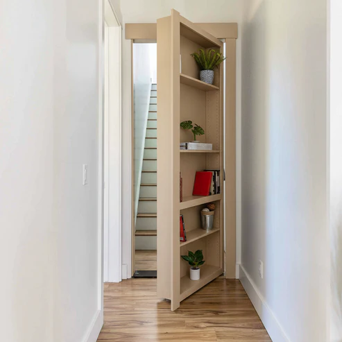 Paint Grade MDF / Unfinished finish variation for Murphy Door® bookcase hidden door in dark wood, partially open to reveal stairs, with decorative plants and books on shelves.